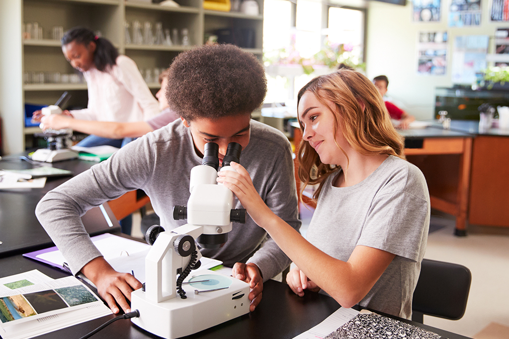 Two high school students in a science lab examine a specimen through a microscope.