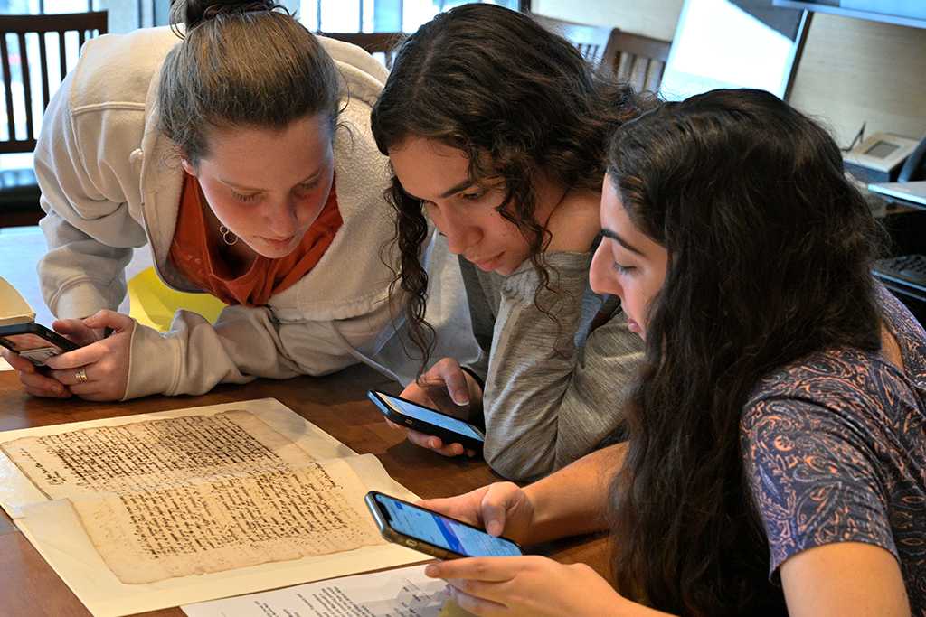 Three students closely review a historic document while referencing information on their cell phones.